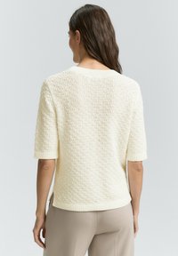 Maglione lavorato a maglia color crema con superficie testurizzata, maniche corte e orlo a coste. Presenta un design semplice e una vestibilità leggermente oversize.