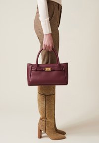 Burgunderfarbene Ledertasche mit strukturierter Form, doppelten Griffen und goldenen Hardware-Akzenten. Getragen zu gemusterten braunen Hosen und hohen Stiefeln.