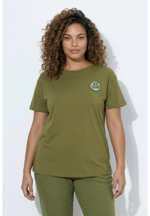 Vrouw met krullend haar die een olijfgroen shirt met korte mouwen en een broek draagt, staand tegen een witte achtergrond met een ontspannen houding.