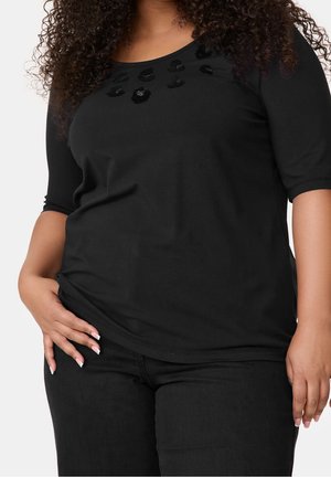 Kiharatukkainen henkilö, joka pukeutunut mustaan paitaan, jossa on kimaltelevia kukkakoristeita kaula-aukon lähellä, ja mustiin housuihin, käsi leväten lantion päällä.