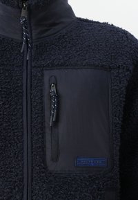 Geacă din fleece de marină, cu un corp texturat, guler înalt și un buzunar din material neted cu închidere prin fermoar și etichetă cu logo.