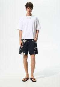 Biele oversized tričko, čierne šortky s bielymi hviezdami a čierne šľapky. Neformálny letný outfit predvedený na modele.