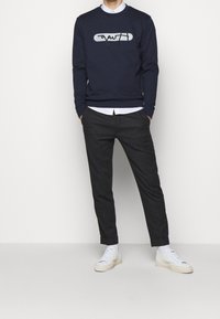 Sudadera azul marino con un diseño gráfico, combinada con pantalones grises ajustados y zapatillas blancas. La camisa debajo es blanca y tiene cuello.