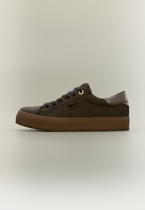Zapatilla deportiva de ante marrón oscuro con cordones negros y suela gruesa marrón, vista desde el lado izquierdo sobre fondo beige.