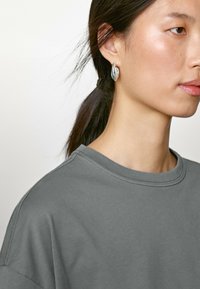 T-shirt en coton gris avec un col rond, doté de manches courtes. Porté avec une boucle d'oreille en argent, en forme de goutte d'eau abstraite.