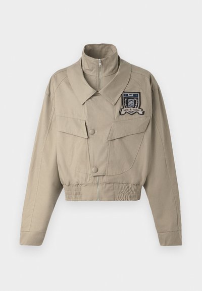 Gekürzte beigen Jacke mit hohem Kragen, vorderem Reißverschluss, zwei großen Brusttaschen und einem gestickten Emblem auf der linken Seite.