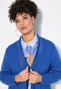 Cárdigan azul de punto con un patrón texturizado, de frente abierto y mangas anchas, superpuesto a una camisa a rayas con un diseño colorido.