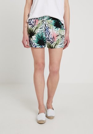 Femme portant un short à imprimé tropical avec des feuilles vertes, noires et roses, un top blanc et des espadrilles blanches, debout devant un fond uni.