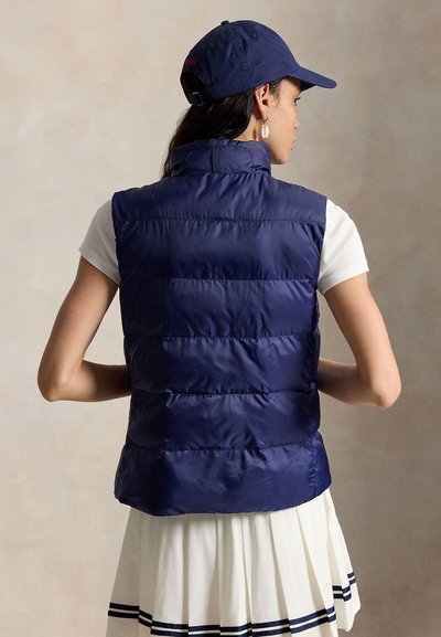 Γυναίκα που φοράει μπλε ναυτικό μπουφάν χωρίς μανίκια, λευκό τοπ με κοντά μανίκια, πλισέ φούστα με μπλε ρίγες, μπλε καπέλο και σκουλαρίκια σε σχήμα κοχυλιού, εμφανιζόμενη από πίσω.