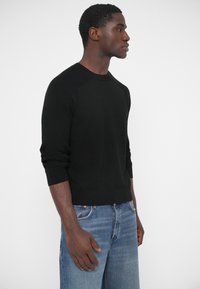 Čierny pletený sveter s rebrovanými detailmi na ramenách, okrúhlym výstrihom a priliehavými manžetami, v kombinácii s modrými džínsami s širokými nohavicami.