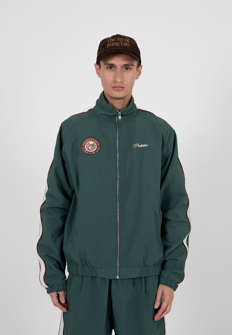 Jeune homme portant une veste zippée vert foncé et un pantalon assorti avec des détails blanc et rouge, ainsi qu'une casquette marron avec un texte doré.