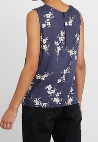 Marinblå ärmlös topp med blommig broderi i vitt och grönt. Har en nyckelhålsöppning bak med dragkedja och en avslappnad passform. Slät textur.