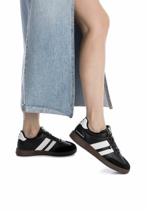 Black sneakers with white stripes, textured rubber sole, smooth leather upper, rounded toe, and contrasting accents. Denim skirt partially visible.