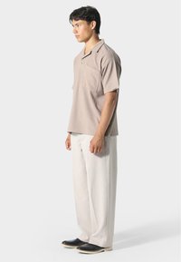 Jasnobrązowa koszulka polo z krótkim rękawem i kieszenią z przodu, w zestawie z luźnymi białymi spodniami i czarnymi butami z białymi podeszwami.