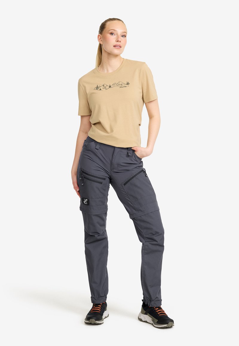 Beige bomull t-shirt med berggrafik. Grå cargobyxor med dragkedjefickor, justerbara ärmslut och textilstruktur. Svarta sneakers med orange detaljer.
