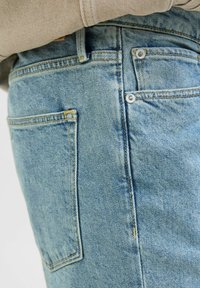 Jeans din denim de un albastru deschis cu cusături vizibile, două buzunare la spate și accesorii din metal argintiu la talie. Textură ușor decolorată.