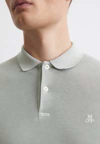 Jasnoszara koszulka polo z teksturowanej tkaniny, z dwoma białymi guzikami i małym białym logo na piersi. Klasyczny krój kołnierzyka.