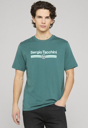 Junger Mann mit lockigem Haar, der ein türkisfarbenes Sergio Tacchini T-Shirt und schwarze Hosen trägt, vor einem hellgrauen Hintergrund stehend.