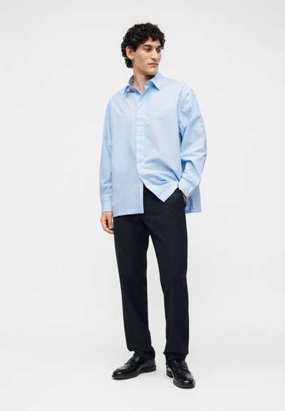 Άνδρας με σγουρά μαλλιά, φορώντας ένα ανοιχτό μπλε φαρδύ πουκάμισο με κουμπιά, μαύρο παντελόνι και μαύρα επίσημα παπούτσια, στέκεται με το ένα χέρι στην τσέπη.