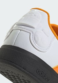 Baskets synthétiques blanches avec des accents orange, semelle en caoutchouc noir texturé, et détail de design moulé au niveau du talon arborant des motifs circulaires.