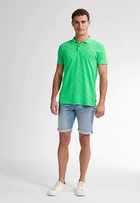 Zielona koszulka polo z małym, powtarzającym się wzorem, w połączeniu z jasnoniebieskimi szortami dżinsowymi. Model nosi białe trampki, stoi na jednolitym tle.