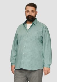 Jasnoniebieska koszula z długim rękawem z małym geometrycznym wzorem, z kołnierzykiem na guziki i jedną kieszenią na piersi. Nosi się ją na białej koszulce.