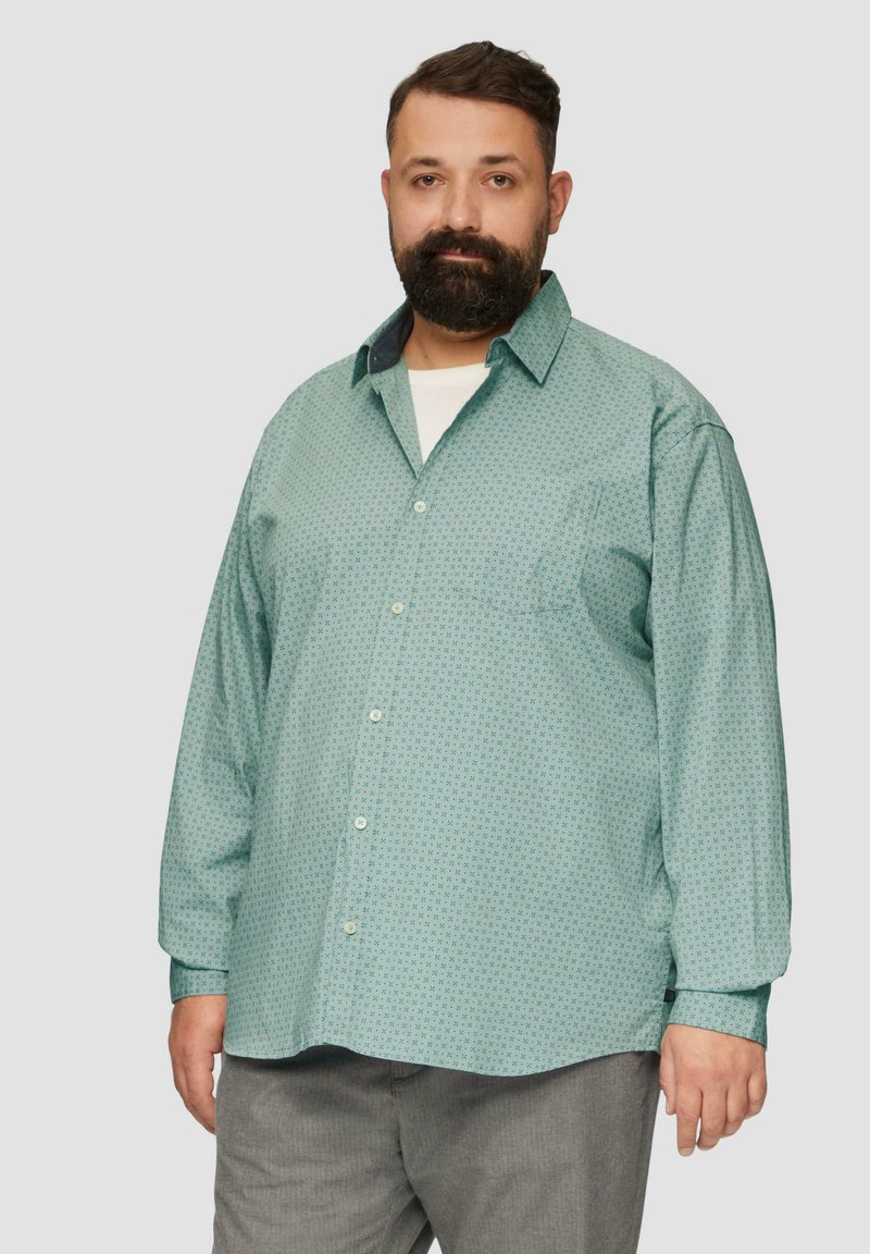 Jasnoniebieska koszula z długim rękawem z małym geometrycznym wzorem, z kołnierzykiem na guziki i jedną kieszenią na piersi. Nosi się ją na białej koszulce.