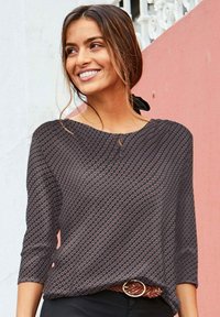 Schwarz gemusterte Bluse mit Dreiviertelärmeln, runder Halslinie und kleinen Aussparungsdetails. Kombiniert mit einem braunen geflochtenen Gürtel.