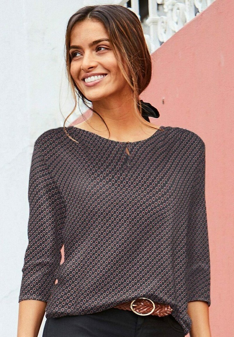 Schwarz gemusterte Bluse mit Dreiviertelärmeln, runder Halslinie und kleinen Aussparungsdetails. Kombiniert mit einem braunen geflochtenen Gürtel.
