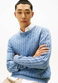 Maglione azzurro chiaro a trecce con collo e polsini a costine, indossato sopra una camicia bianca con colletto. Il modello ha le braccia incrociate, su uno sfondo neutro.