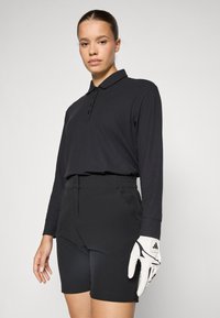Black long-sleeve polo with a button placket, paired with black shorts. The model wears a white golf glove featuring black accents.