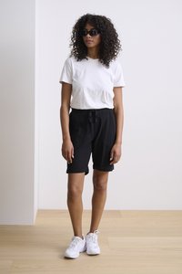 T-shirt blanc, shorts noirs à cordon de serrage et baskets blanches. Possède des cheveux bouclés et des lunettes de soleil noires. Design simple et décontracté.