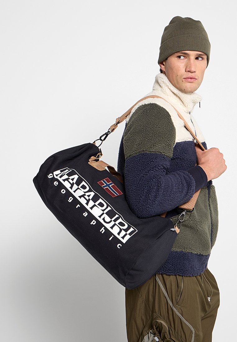 Man in een kleurverloop fleecejack en beanie, met een zwarte Napapijri duffelbag met een Noorse vlag patch op de riem.