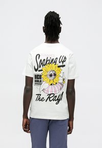 Valkoinen puuvillainen t-paita, jossa on värikäs aurinkohahmo aurinkolaseissa, teksti kuuluu "Sunnie’s Soaking Up The Rays!"