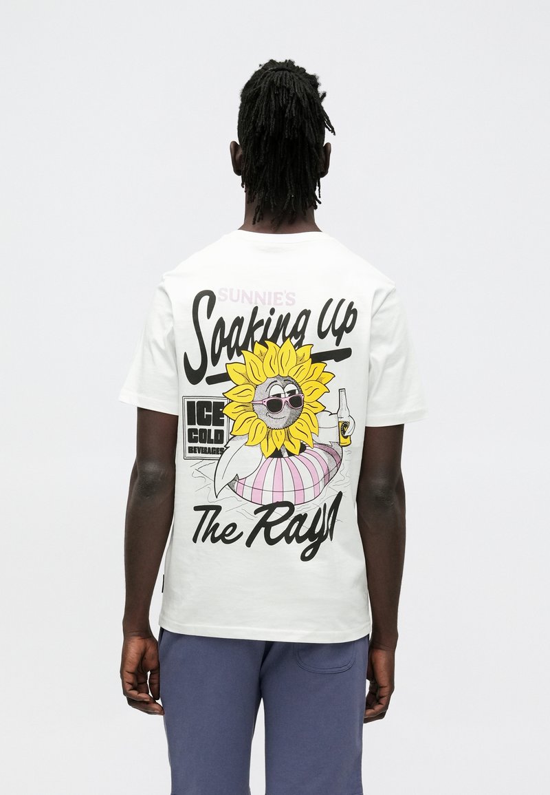 Valkoinen puuvillainen t-paita, jossa on värikäs aurinkohahmo aurinkolaseissa, teksti kuuluu "Sunnie’s Soaking Up The Rays!"