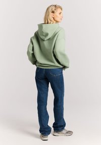 Groene oversized hoodie met een capuchon, gedragen met blauwe rechtlopende jeans en grijze sneakers. De stof lijkt zacht en heeft een casual pasvorm.