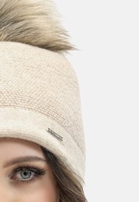 Bonnet en tricot couleur beige clair avec un design texturé, doté d'un grand pompon en fausse fourrure et d'une étiquette de marque en argent sur le côté.