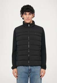 Chaleco acolchado negro con cuello alto, cierre de cremallera y bolsillos laterales. Llevado sobre una camiseta de manga larga negra, combinado con jeans de mezclilla azul claro.