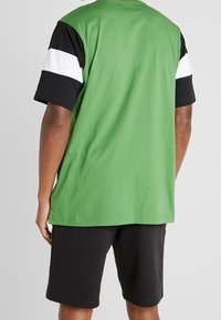 Camiseta verde de manga corta con detalles de rayas negras y blancas en las mangas, combinada con pantalones cortos negros, la tela parece suave y casual.