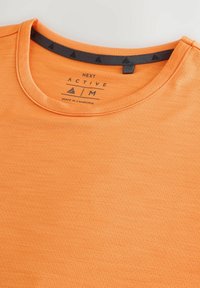 Aktiv t-shirt i lys orange, med rund halsudskæring, en struktureret stof og et gråt logo label med trekantede detaljer på den indvendige krave.