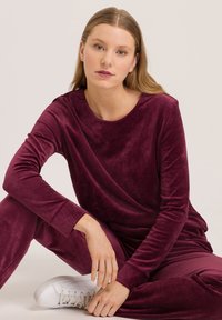 Conjunto de loungewear de terciopelo burdeos con mangas largas y cuello redondo. El modelo lleva zapatillas blancas. Textura suave, ajuste relajado.