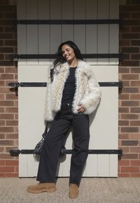 Giacca in faux fur color crema con macchie marroni, abbinata a jeans neri a gamba larga e stivali marroni con lacci. Gli accessori includono una borsa nera.