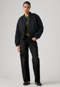 Μαύρο μπουφάν μπότερ με εφαρμοστή γραμμή, διαθέτει ελαστικά μανίκια, φερμουάρ μπροστά και πλαϊνές τσέπες. Φορεμένο πάνω από ένα crop top ελιά πράσινου χρώματος και μαύρα τζιν.