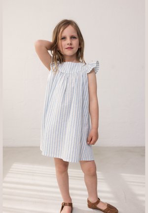 Jeune fille aux cheveux blonds portant une robe blanche sans manches à rayures bleues et des sandales marron, debout avec un bras levé derrière la tête.