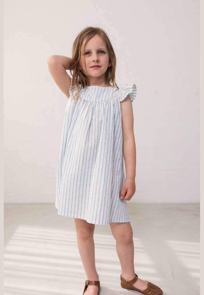 Νεαρή κοπέλα με ξανθά μαλλιά που φοράει λευκό αμάνικο φόρεμα με μπλε ρίγες και καφέ σανδάλια, στέκεται με το ένα χέρι ανασηκωμένο πίσω από το κεφάλι της.