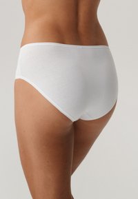 Kobieta w prostych białych bawełnianych majtkach widziana z tyłu na neutralnym tle.