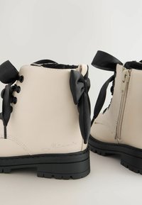 Beige Stiefeletten mit schwarzen Satinschleifen, Schnürung und seitlichem Reißverschluss, strapazierfähige Gummisohle und dezente geprägte Details an der Seite.
