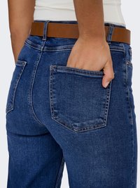 Tamsiai mėlyni džinsai iš džinso audinio su tiesiu kirpimu, kuriuose yra dvi galinės kišenės ir rudas odinis diržas. Audinys turi lygų paviršių.