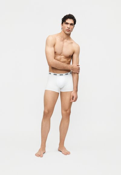 Jeune homme debout, pieds nus, portant un boxer blanc GCDS, torse nu, avec un bras croisé sur sa poitrine, sur un fond clair uni.