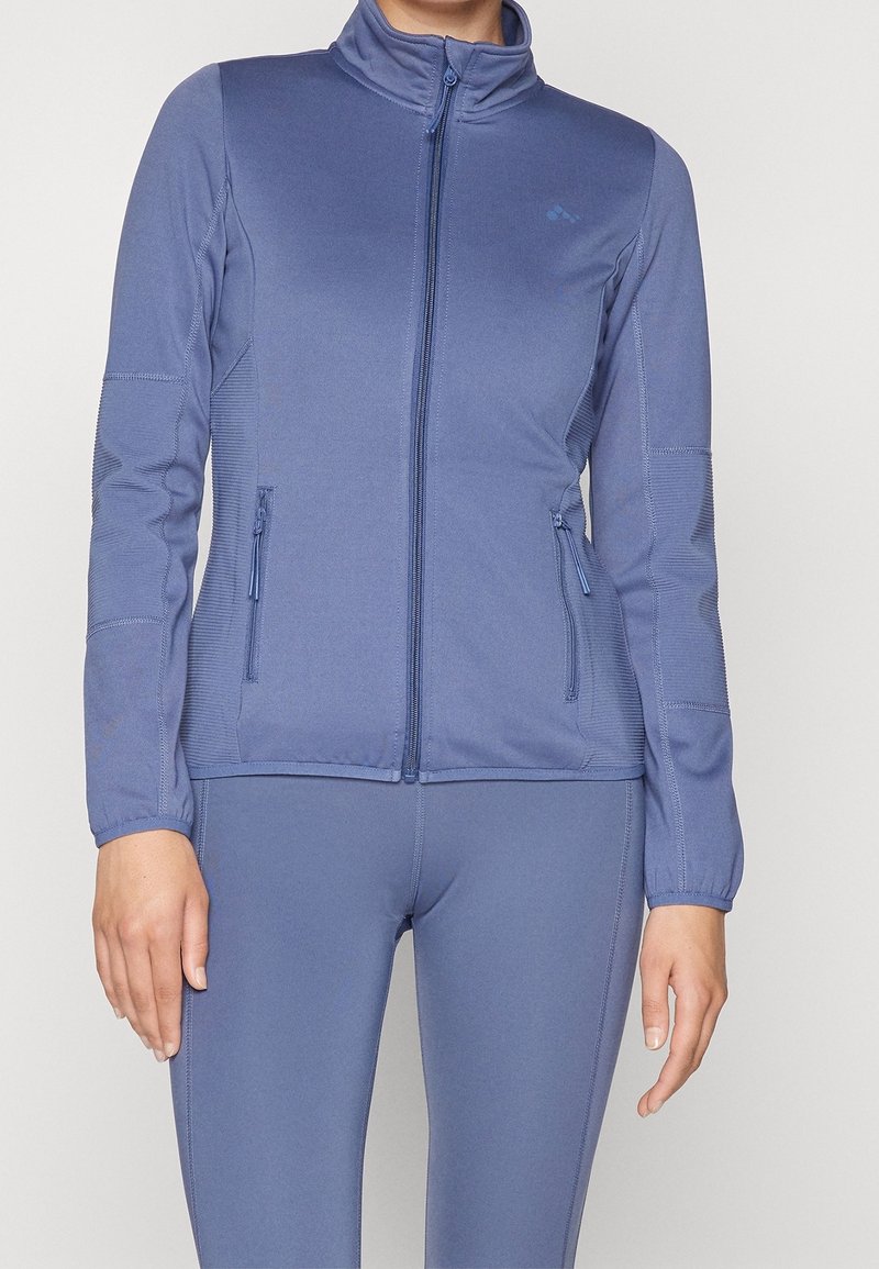 Blå zip-jacka i slät tyg med ståkrage, två framfickor och texturerade detaljer på ärmarna.